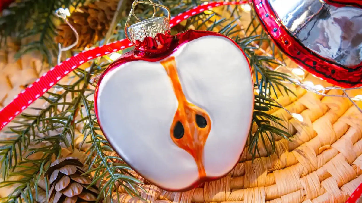A sliced apple blown glass Christmas ornament, on a woven mat with pine branches and a red ribbon.