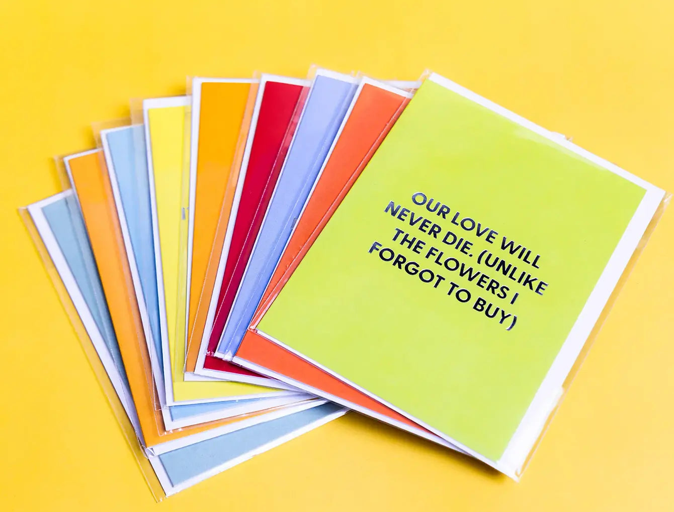 A fanned-out set of colorful greeting cards on a solid yellow background. The front card is lime green with black text reading 'OUR LOVE WILL NEVER DIE (UNLIKE THE FLOWERS I FORGOT TO BUY)'.