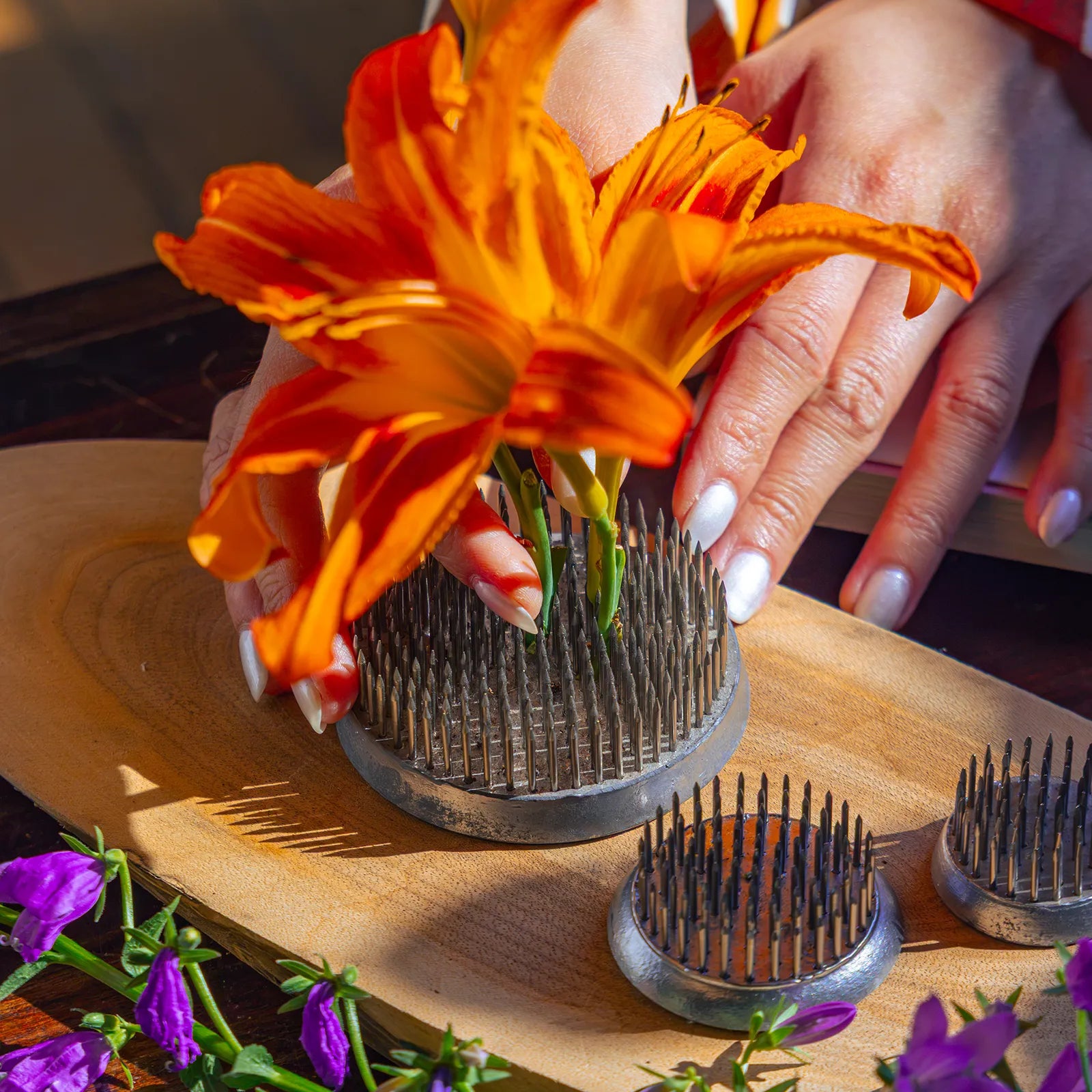 A person's hand pressing fresh orange lilies onto a metal flower frogs on a wooden board.