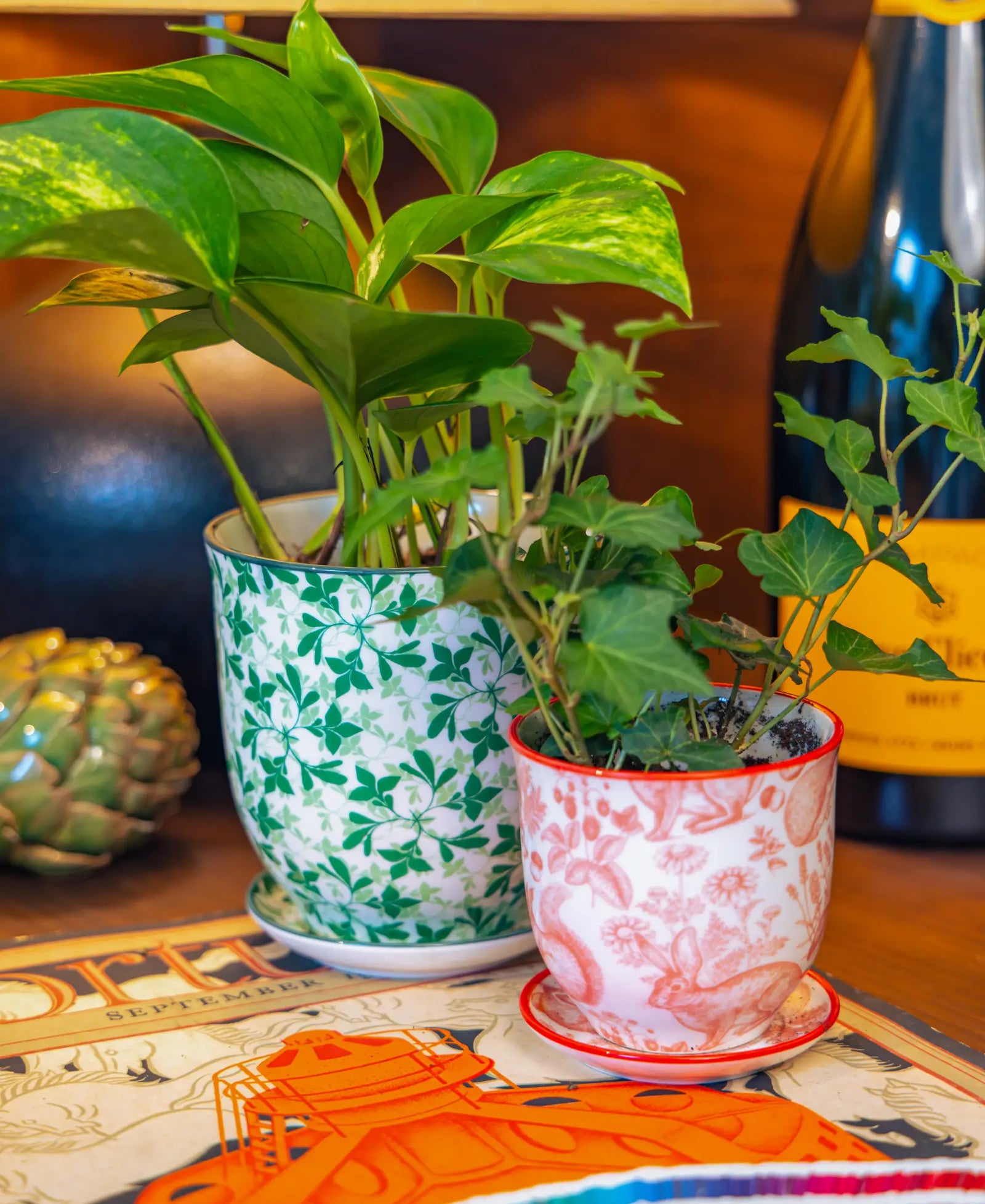 Chive Liberté planters in green leaves and red squirrels prints, styled with a golden pothos and ivy on a wood surface.
