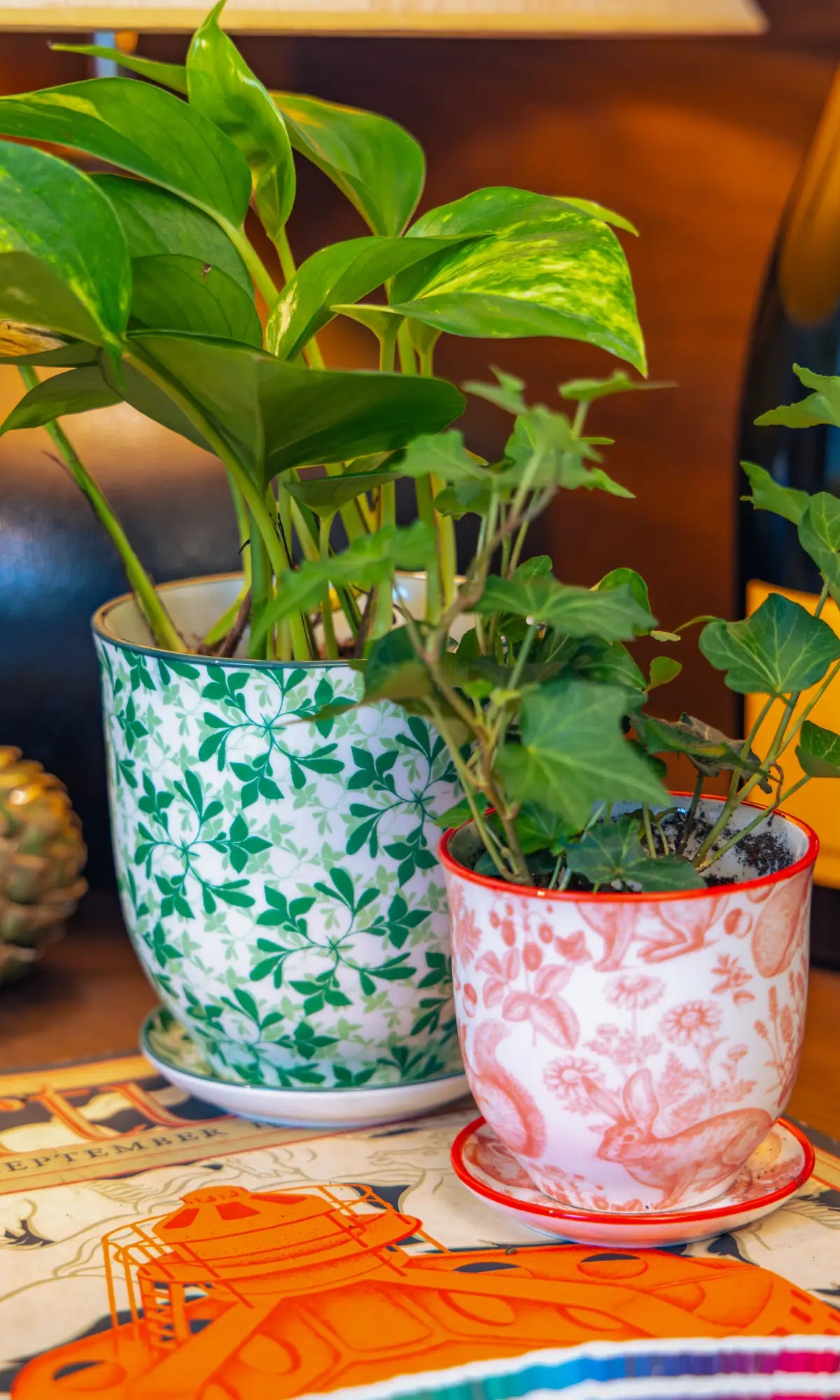 Chive Liberté planters in green leaves and red squirrels prints, styled with a golden pothos and ivy on a wood surface.