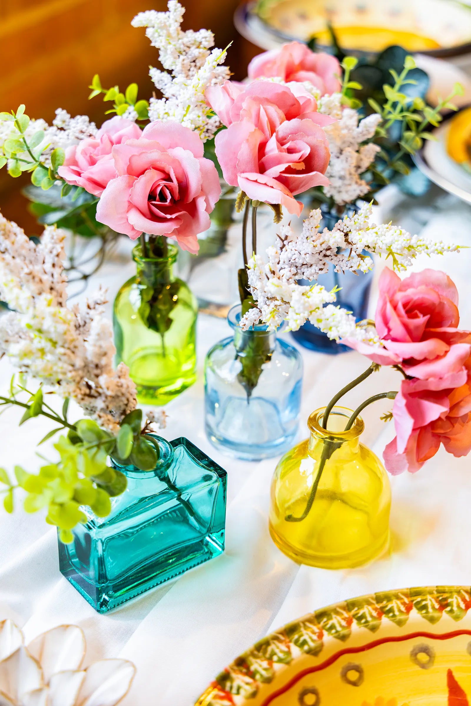 A wedding tabletop arrangement with six Chive Loft glass bud vases in teal, green, light blue, dark blue, clear, and yellow holding pink and white flowers.