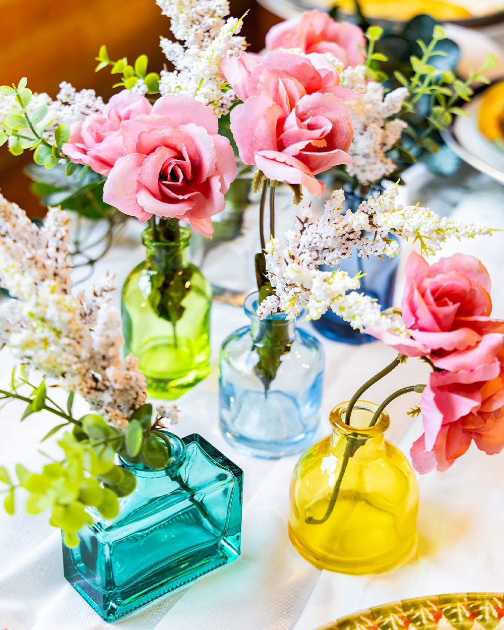 A wedding tabletop arrangement with six Chive Loft glass bud vases in teal, green, light blue, dark blue, clear, and yellow holding pink and white flowers.