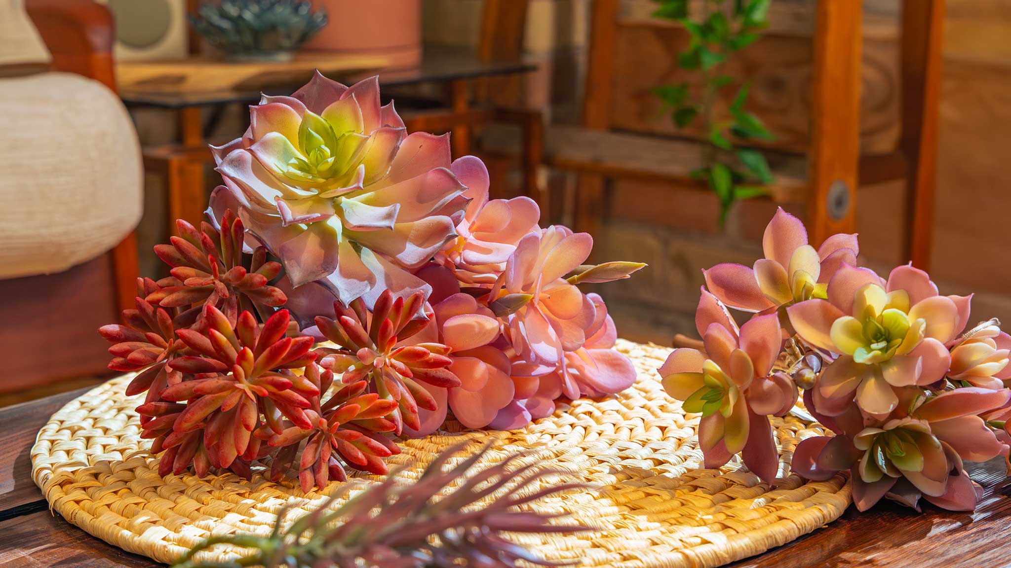 A cluster of Chive realistic pink-yellow faux succulent echeveria stems arranged on a woven round mat on a wood table.