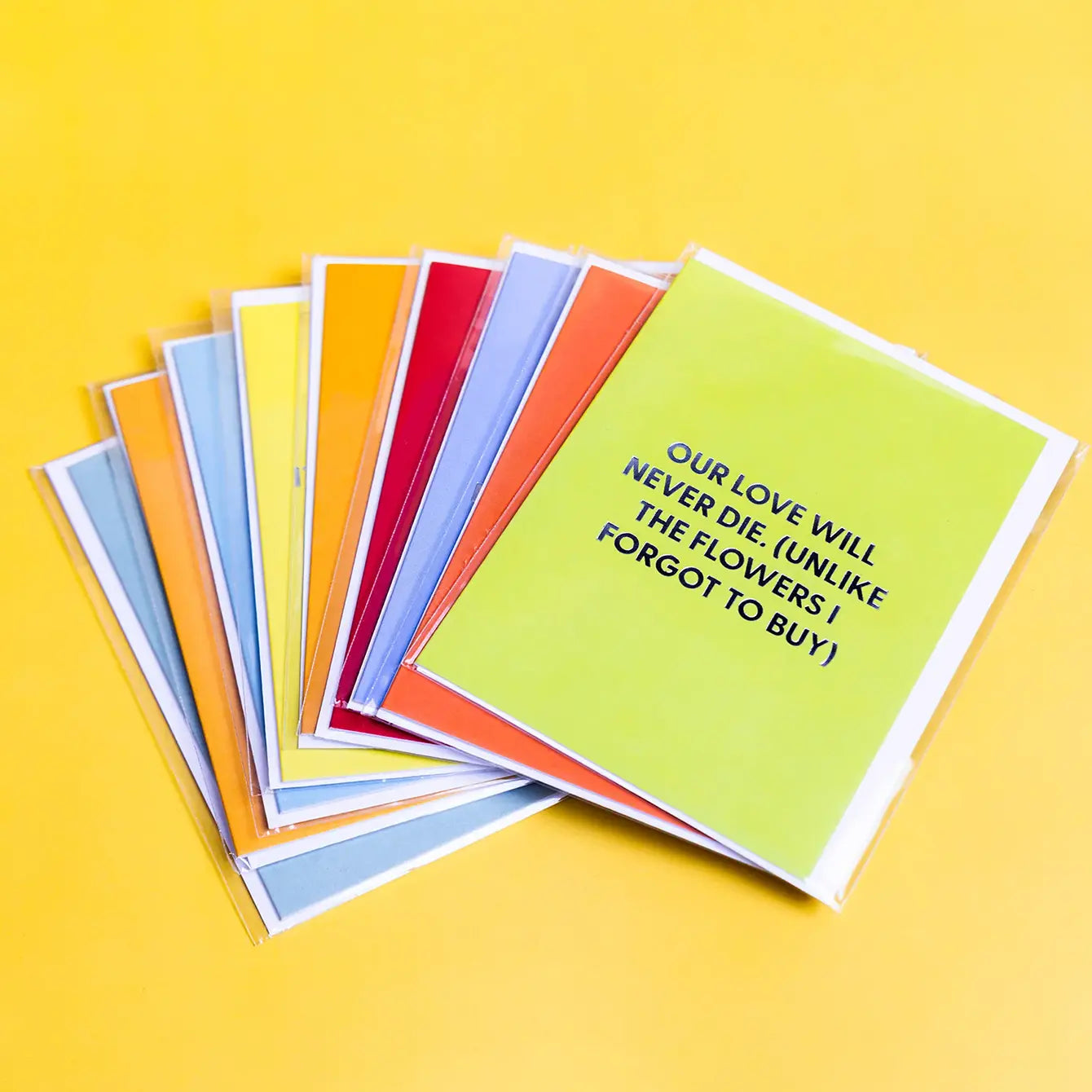 A fanned-out set of colorful greeting cards on a solid yellow background. The front card is lime green with black text reading 'OUR LOVE WILL NEVER DIE (UNLIKE THE FLOWERS I FORGOT TO BUY)'.