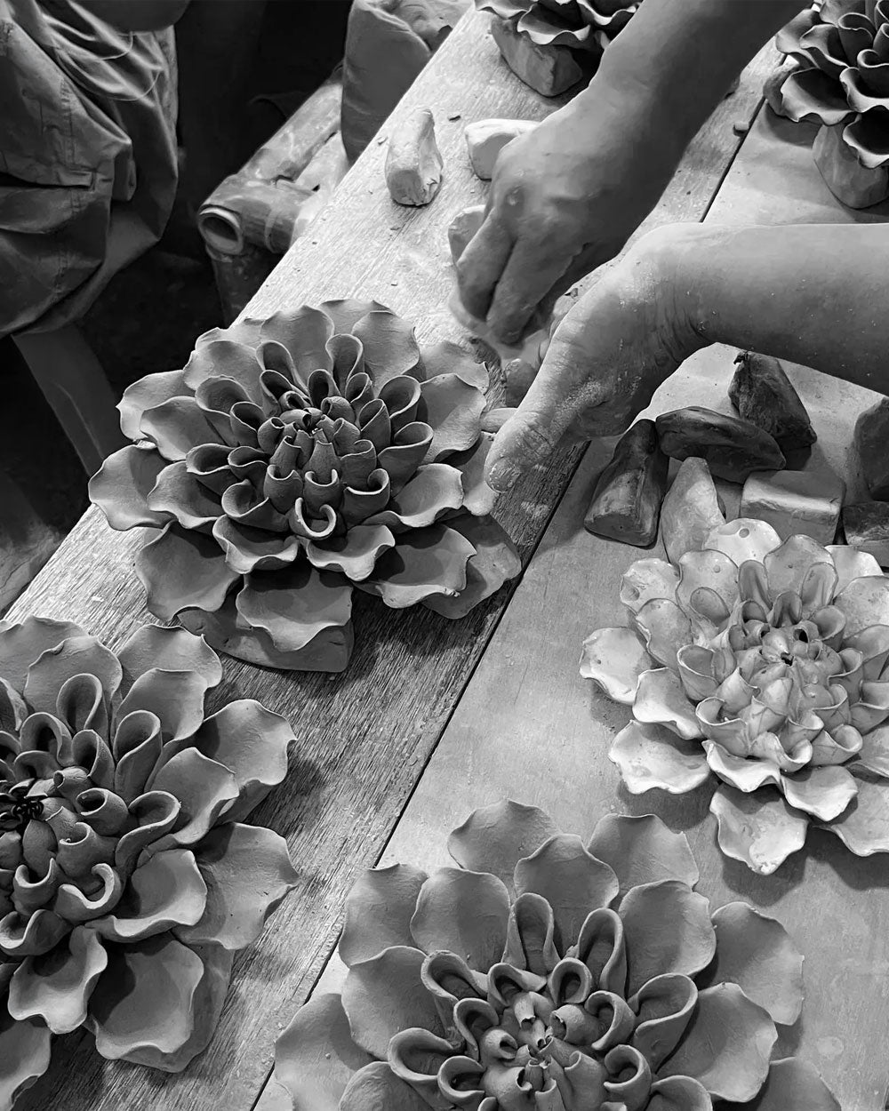 Black and white photo of a craftsman's hands shaping a detailed clay flower on a wooden workbench surrounded by other clay flowers. | Chive