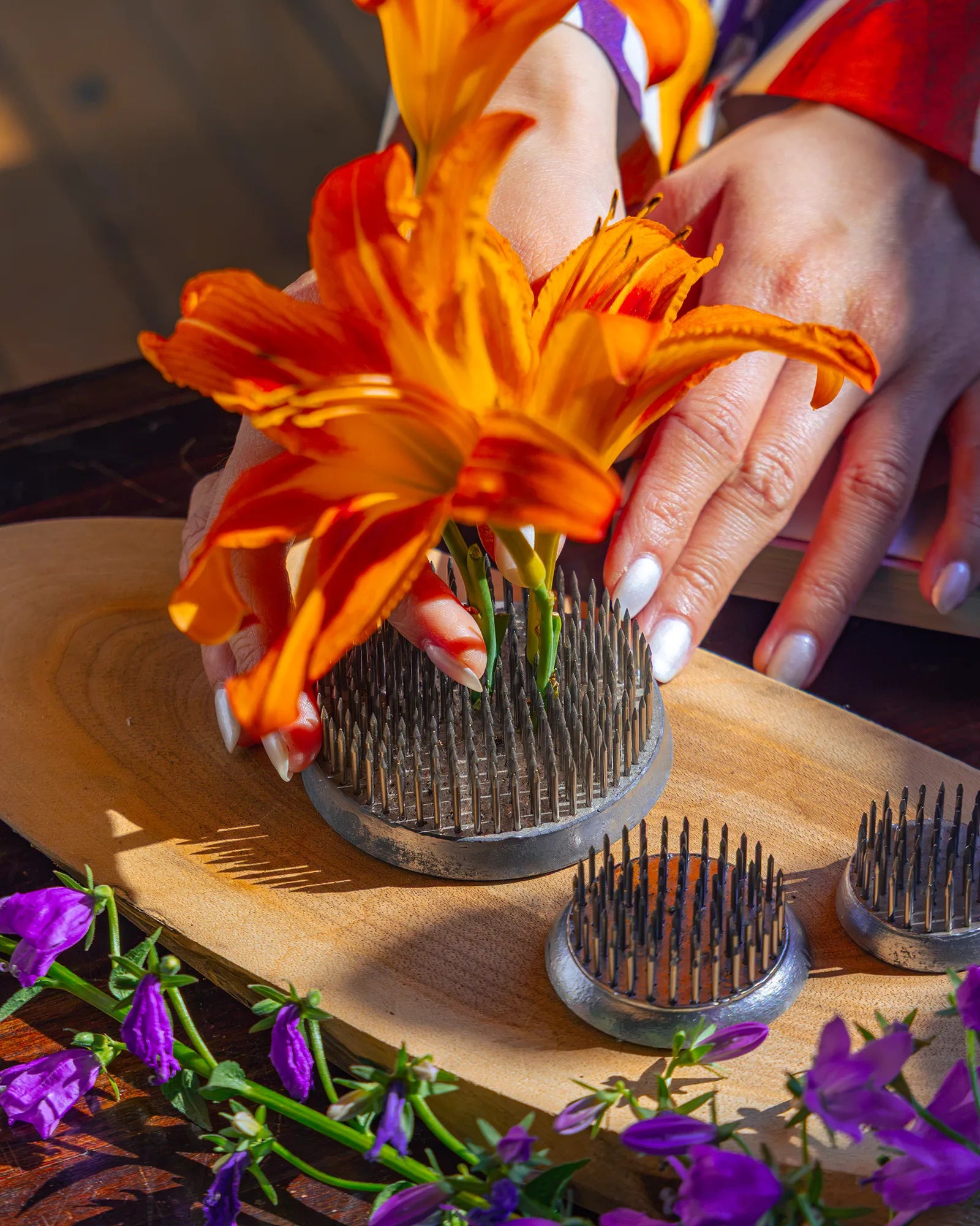 A person's hand pressing fresh orange lilies onto a metal flower frogs on a wooden board.