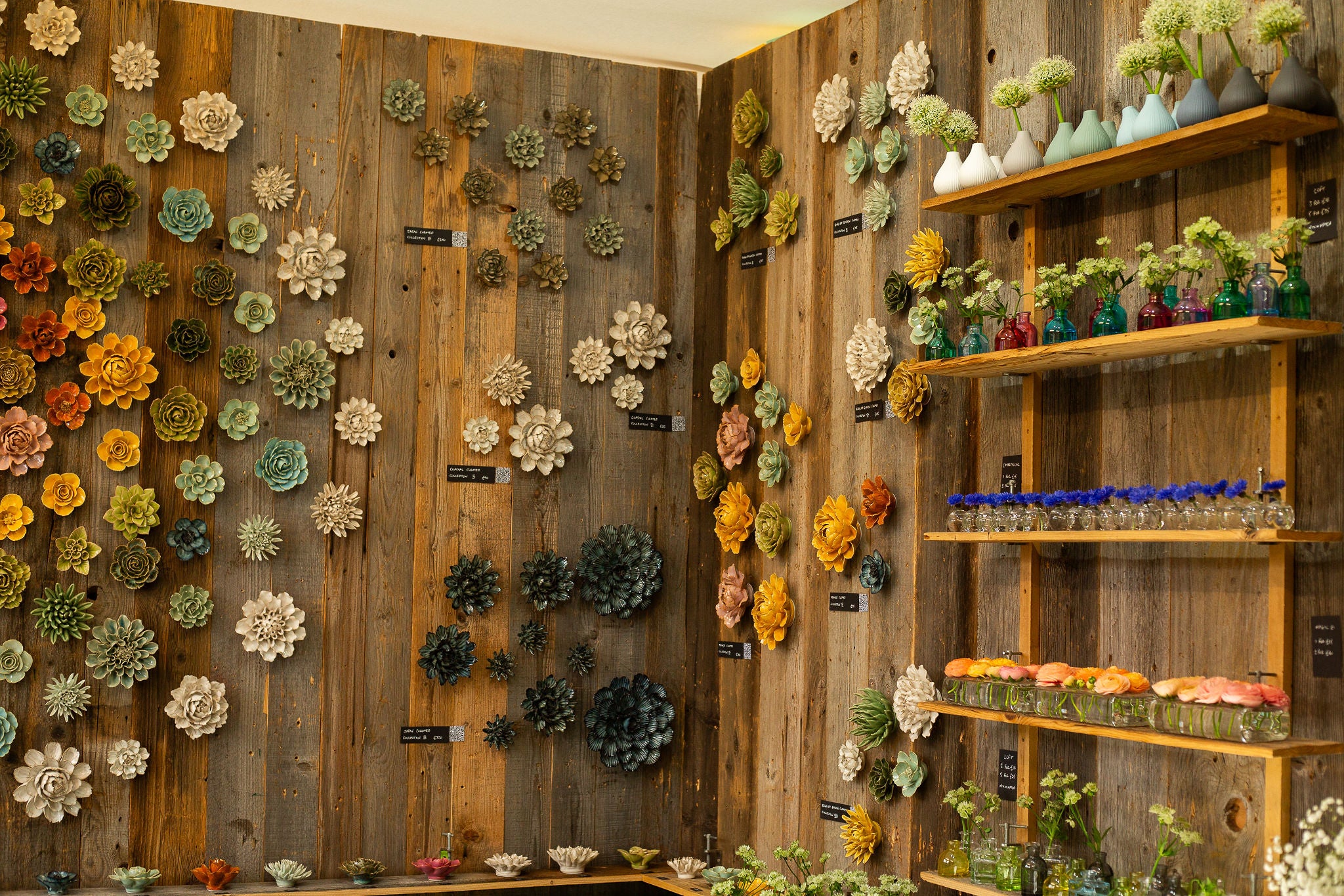 Chive Studio ceramic wall flowers displayed in a circular arrangement on rough wood walls at RHS Chelsea Flower Show booth with ceramic and glass vases on shelves — Chive Studio
