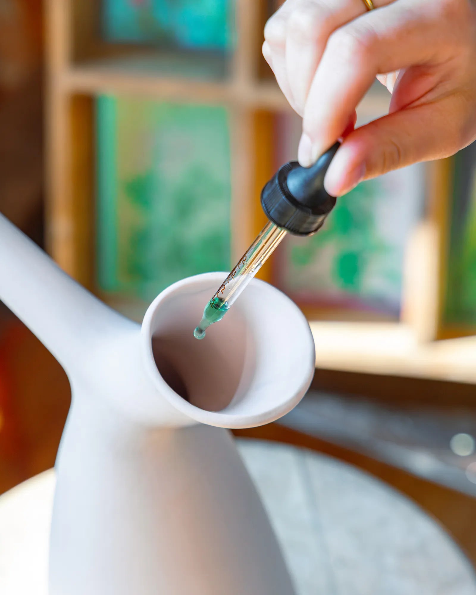 A close-up of a hand holding a Chive Verte Rx plant vitamin glass dropper above a white ceramic watering jug.