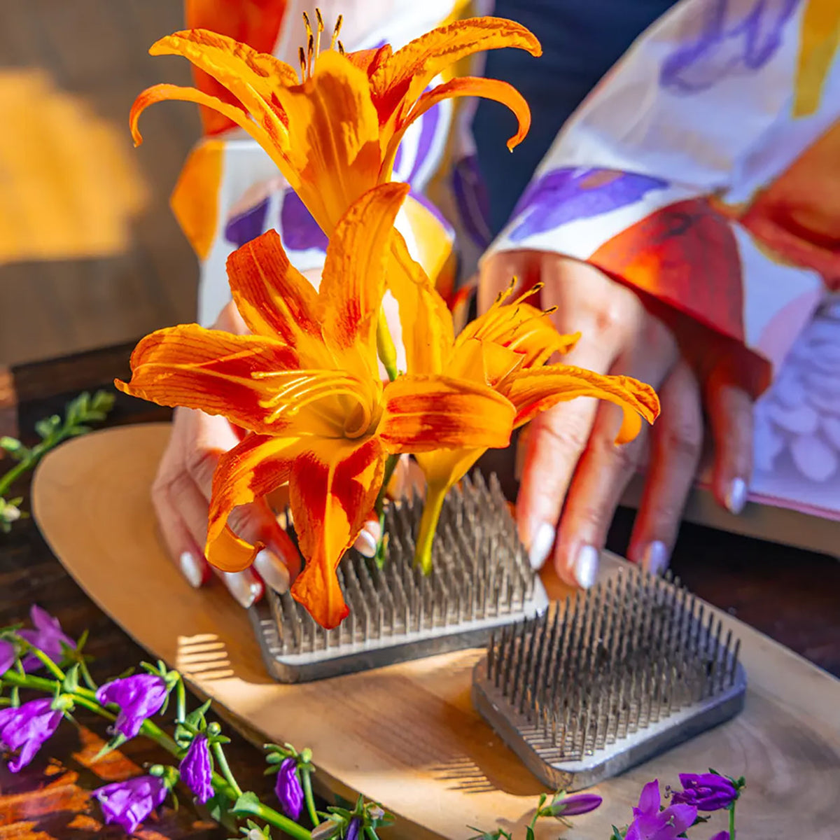 A person's hand pressing fresh orange lilies onto two metal flower frogs on a wooden board.