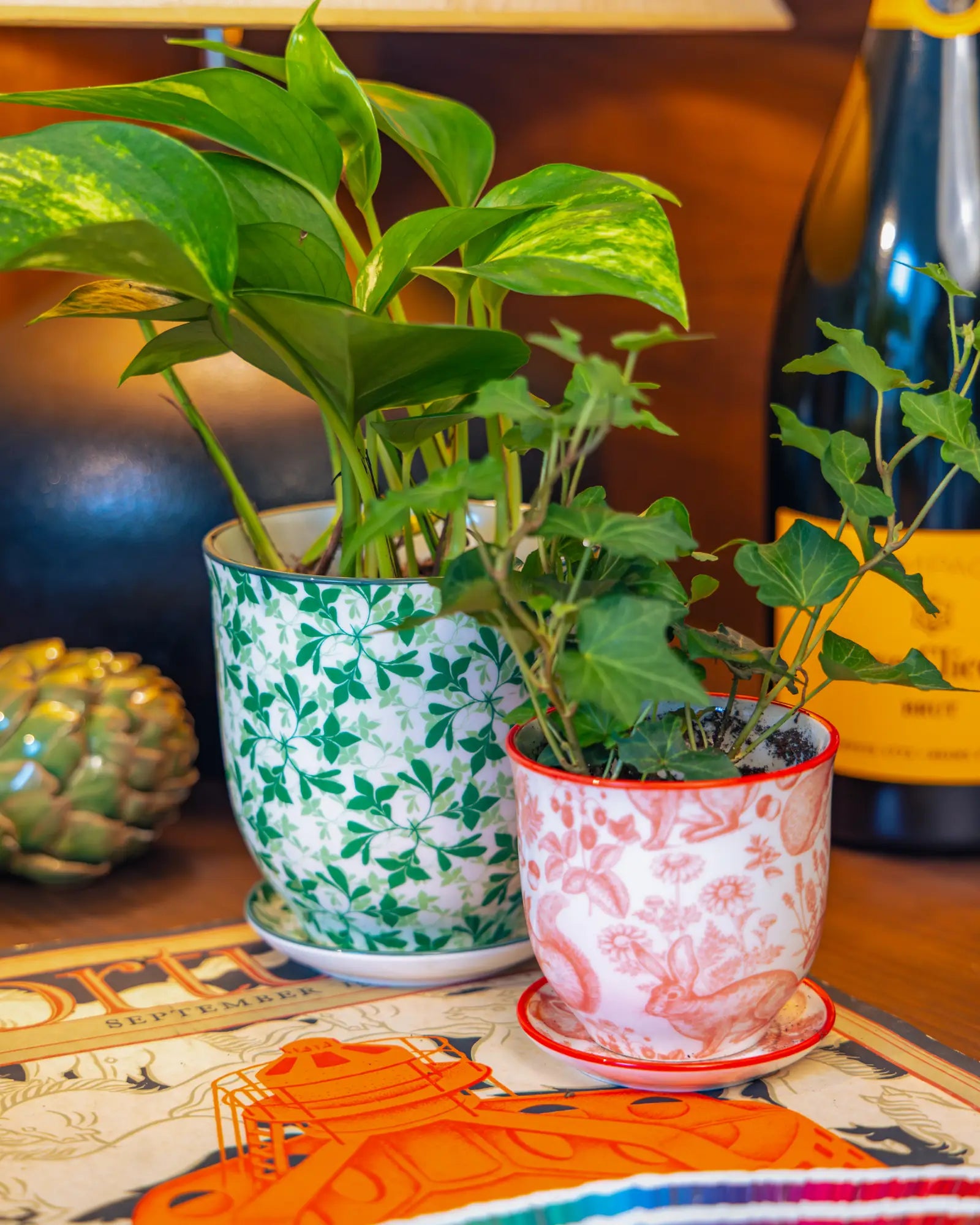 Chive Liberté planters in green leaves and red squirrels prints, styled with a golden pothos and ivy on a wood surface.