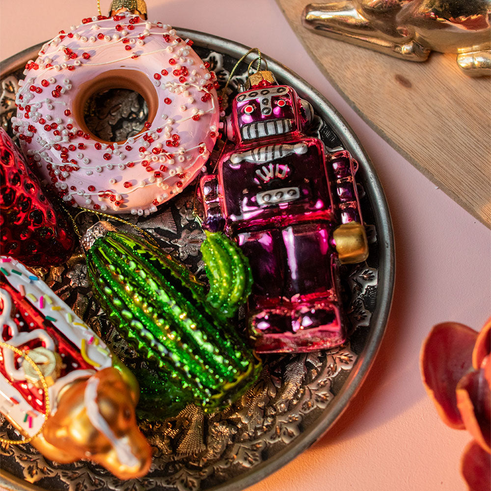 A close-up of a decorative tray holding Chive's colorful holiday ornaments, including a pink donut with sprinkles, a green cactus, a metallic magenta robot, and other ornaments on a pink surface.